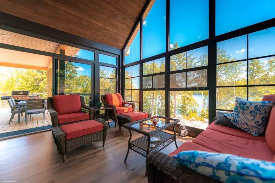 Modern sunroom with black-framed floor-to-ceiling windows and wicker seating with red cushions overlooking a lake