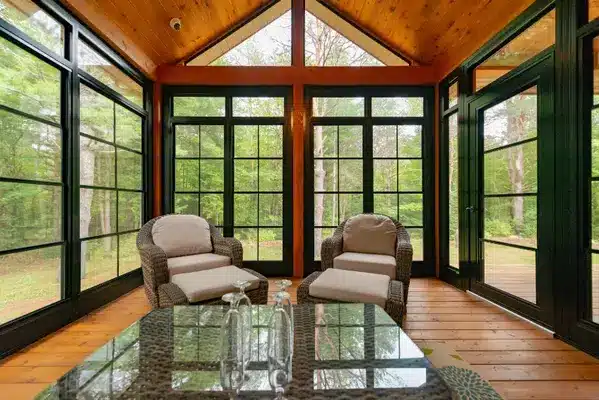 A rustic sunroom with a vaulted wood ceiling, black-framed windows, wicker furniture, and a floral area rug overlooking a forest.
